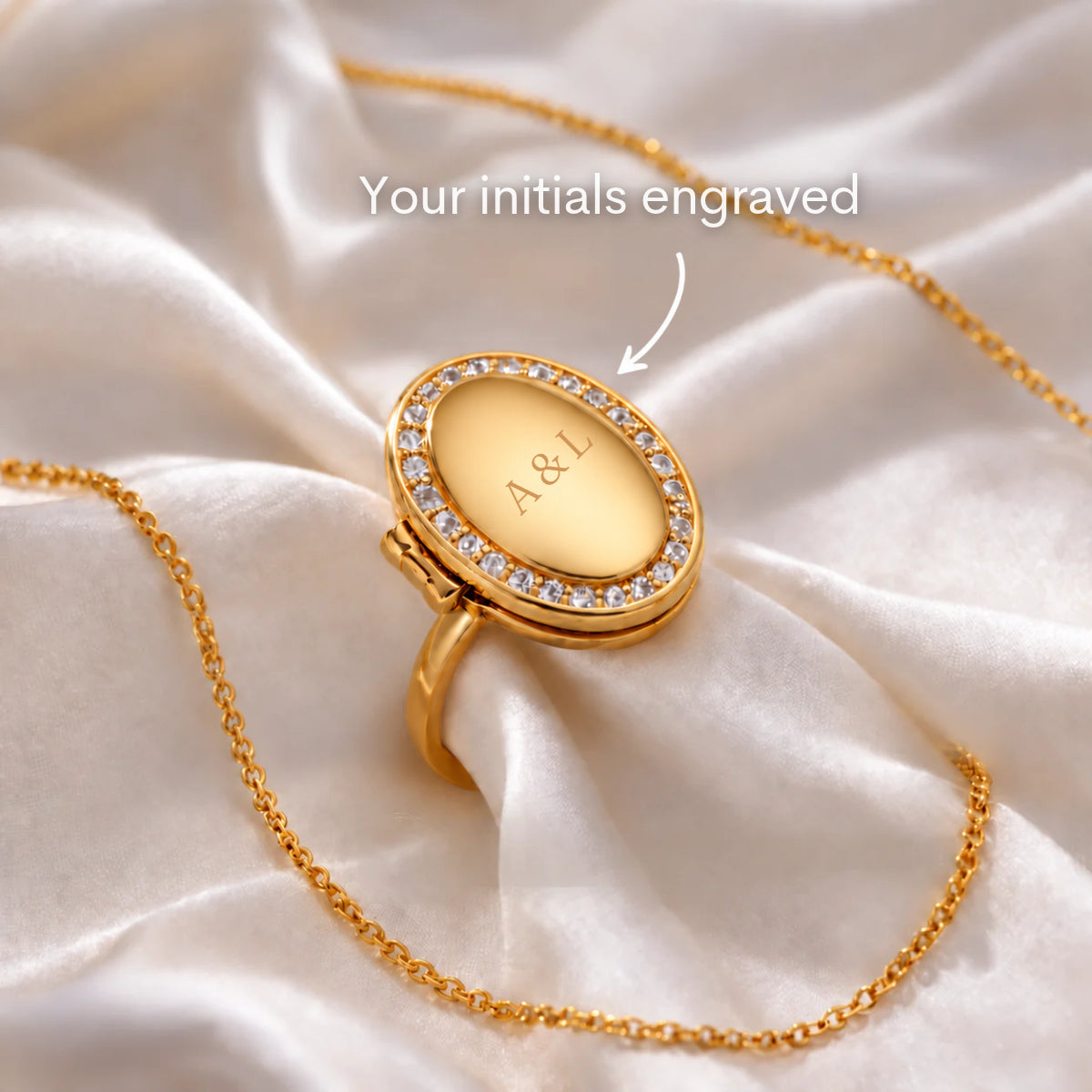 Oval Locket Ring in gold, closed, with initials engraved on the front of the oval locket, framed by a halo of small clear stones.